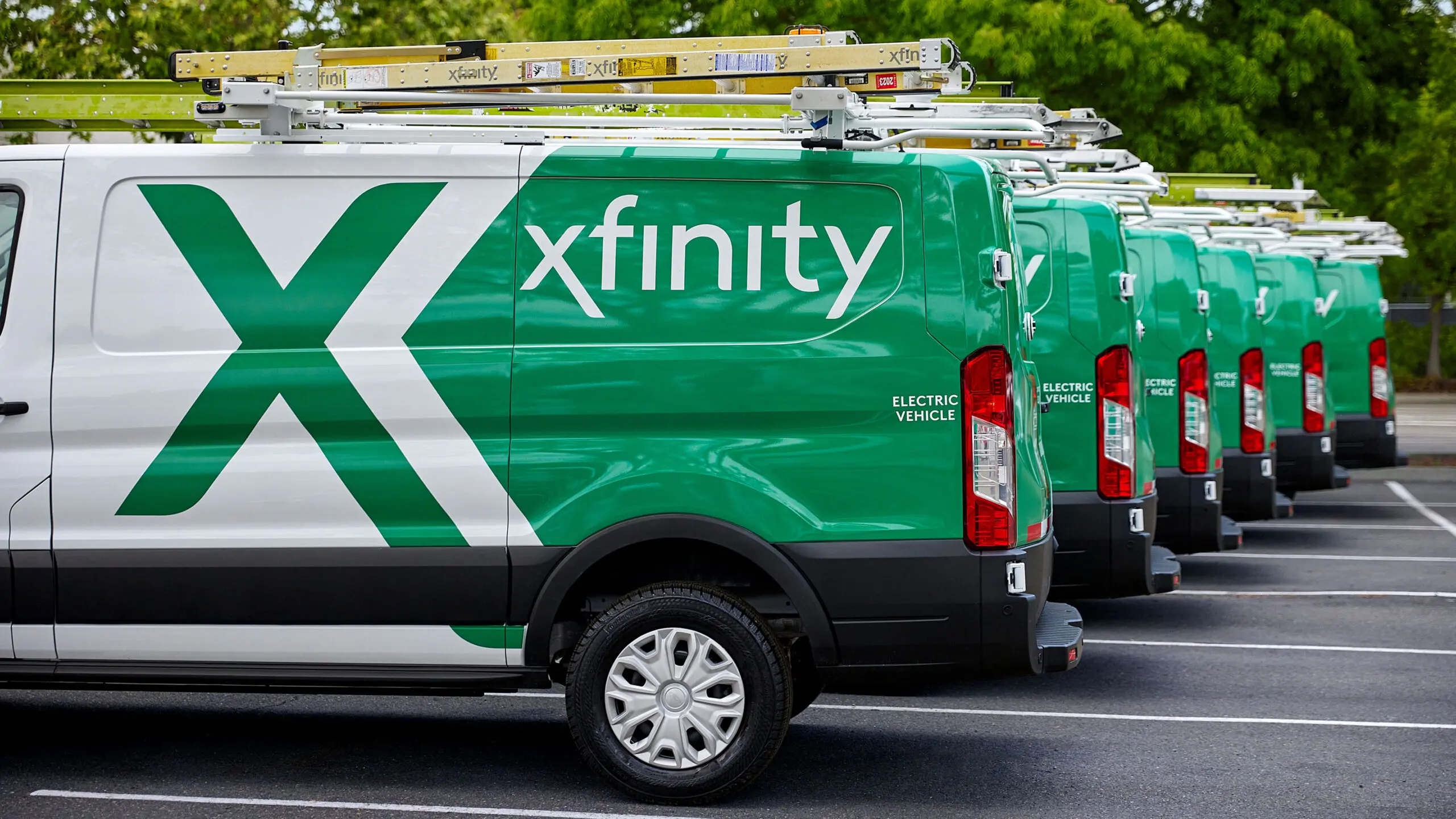 A row of Xfinity service vans parked in a parking lot. The vans are green and white in color with the Xfinity logo prominently displayed on the sides. They have ladder racks mounted on top and are labeled as "ELECTRIC VEHICLE." The image shows multiple identical vans lined up in succession, creating a repeating pattern.