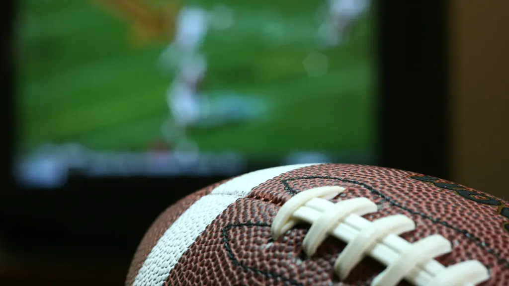 Close up of a football with a television playing a football game in the background.