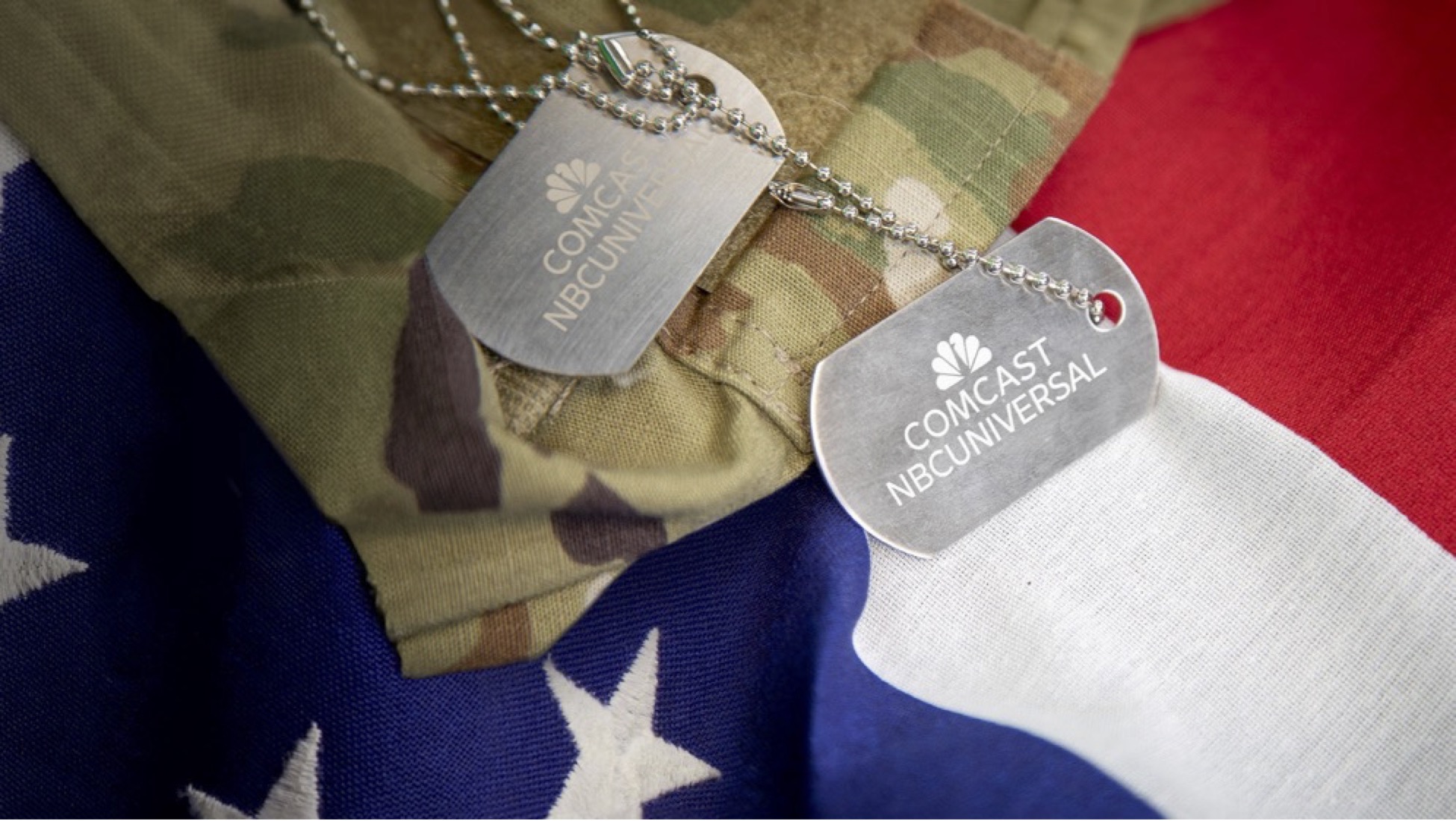 Military-style dog tags with the logos of 'Comcast NBCUniversal' and 'Comcast NBCUniversal' lying on a background of camouflage fabric and an American flag. The dog tags are silver/metallic in color with engraved white logos.